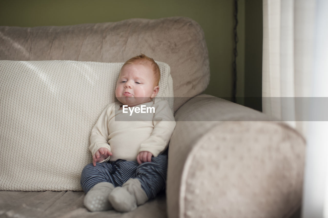 Cute baby boy frowning while sitting on sofa at home