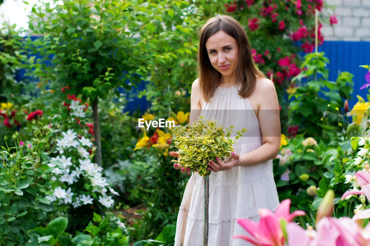 flower, flowering plant, plant, women, one person, adult, floristry, nature, smiling, freshness, growth, beauty in nature, happiness, young adult, long hair, gardening, hairstyle, portrait, garden, lifestyles, emotion, standing, front view, brown hair, holding, summer, outdoors, female, cheerful, day, floral design, bouquet, looking at camera, flower arrangement, three quarter length, green, dress, fashion, leisure activity, fragility, ornamental garden, looking, business, spring, flowerbed, yellow, bride, rose, casual clothing, botany