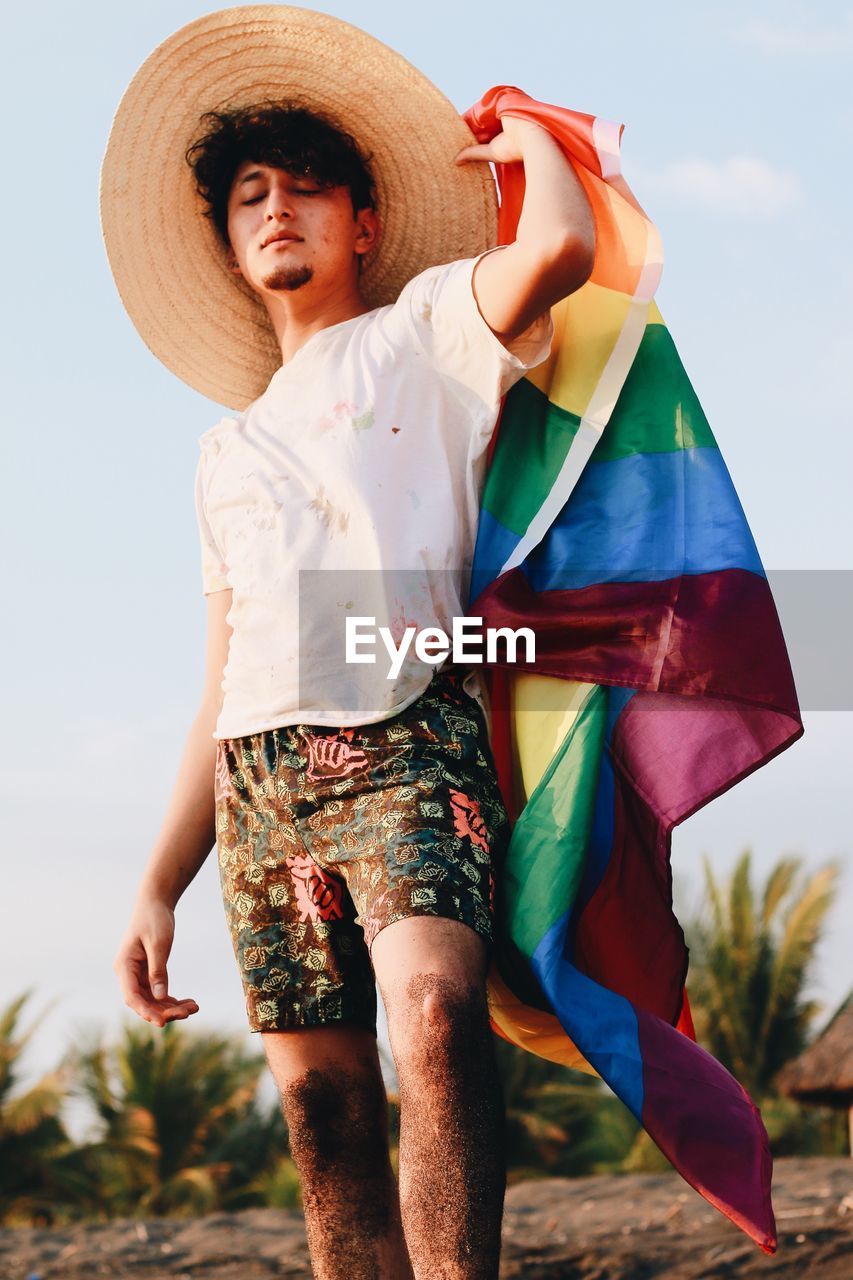 Young man holding rainbow flag while standing at beach