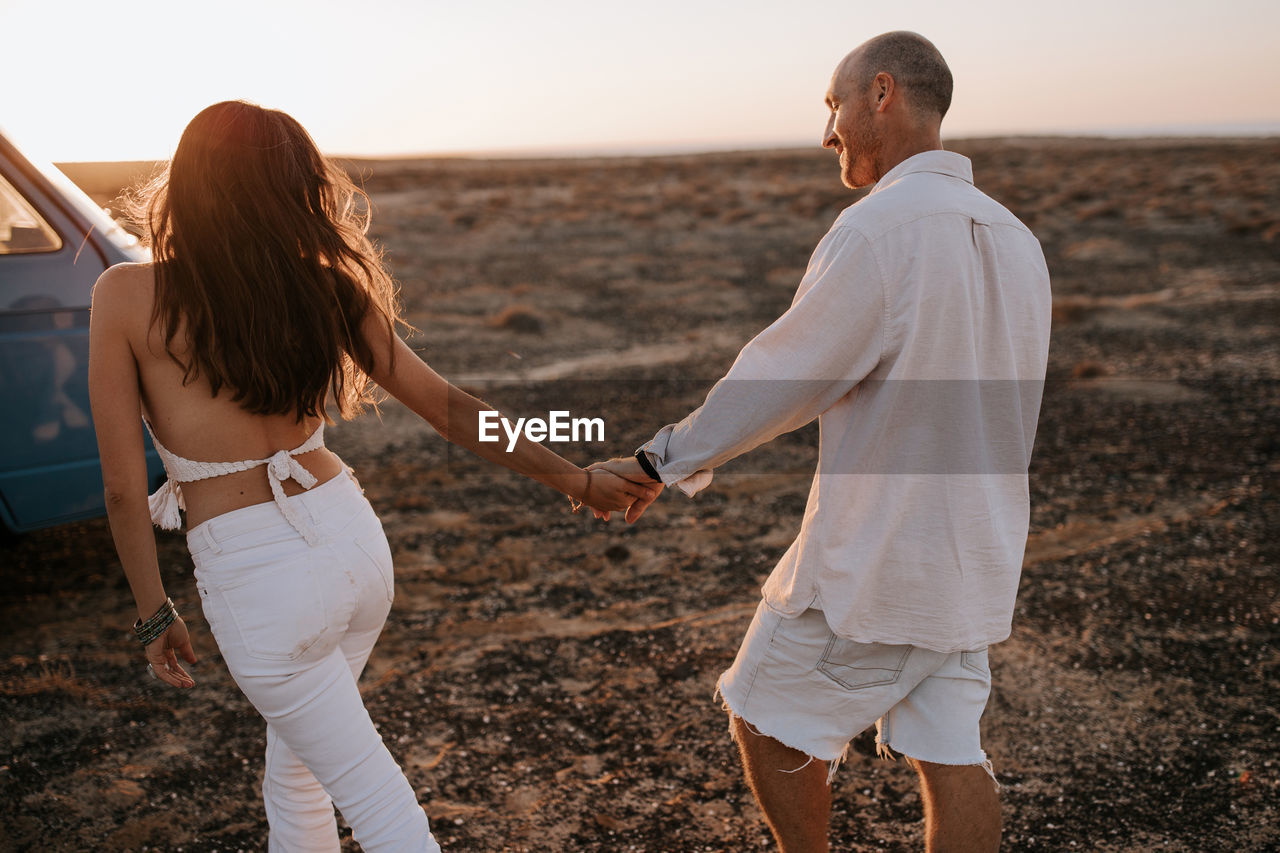 Back view of couple of travelers holding hands while walking in savanna at sunset during summer vacation