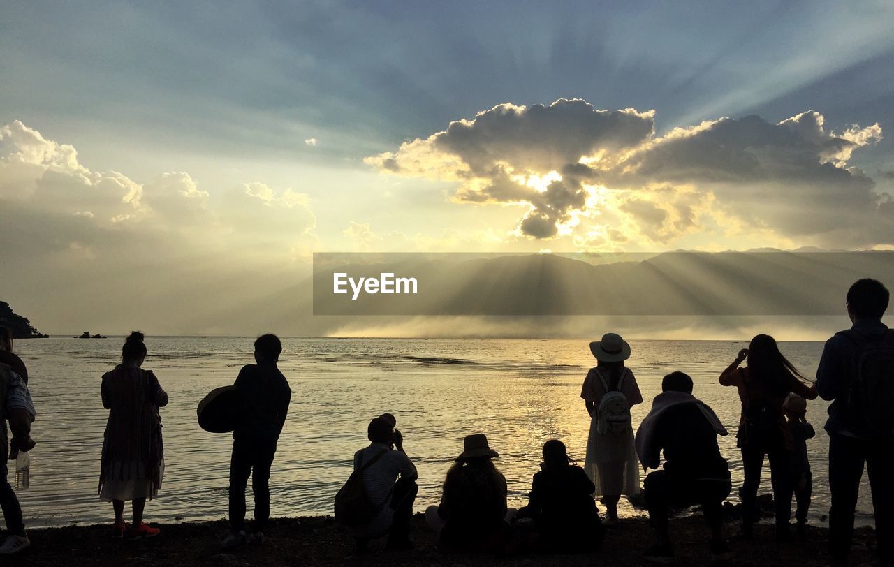 Rear view of people at beach against cloudy sky during sunset