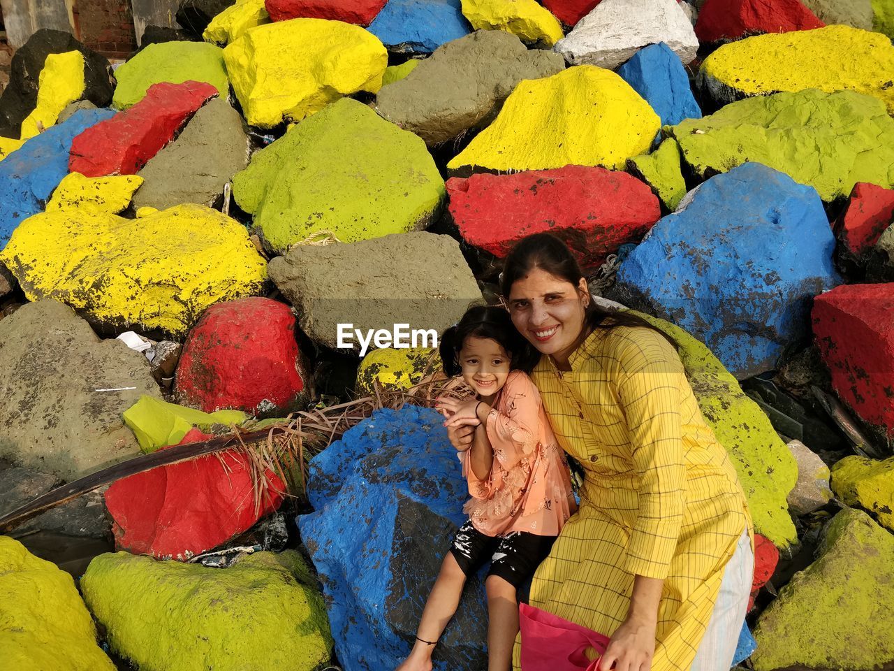 Portrait of a smiling young woman with daughter sitting on colorful rocks at beach