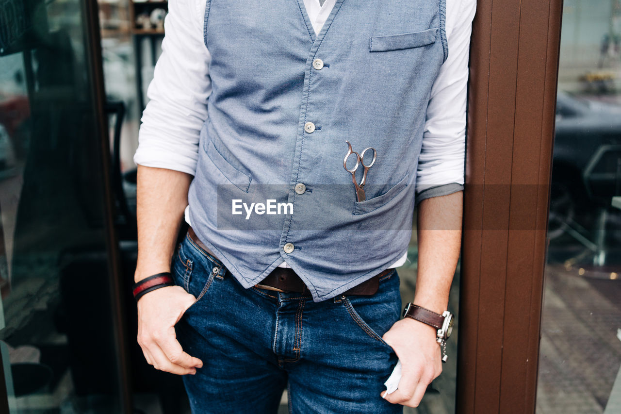 Cropped unrecognizable adult bearded male beauty master with hands in pockets at door of barbershop