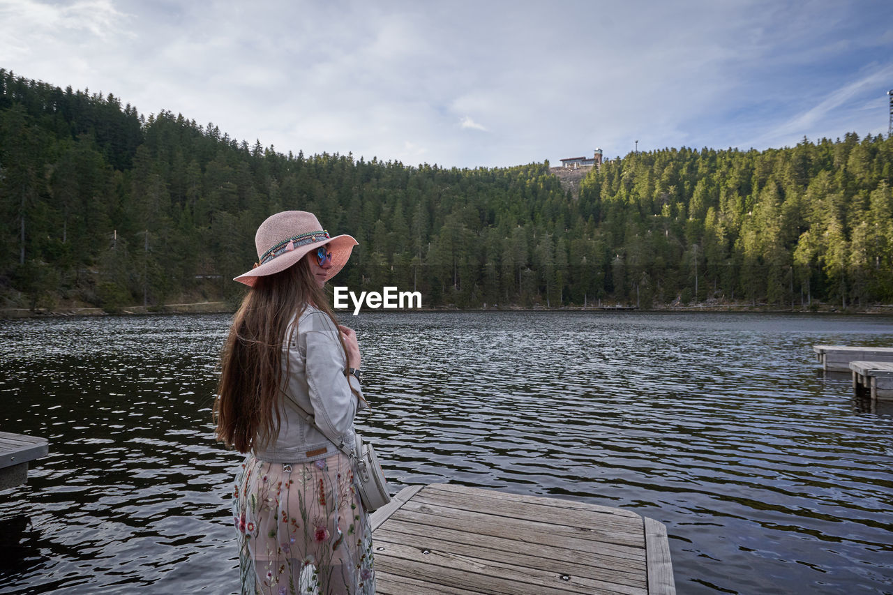 Woman wearing hat while standing on pier by lake against sky