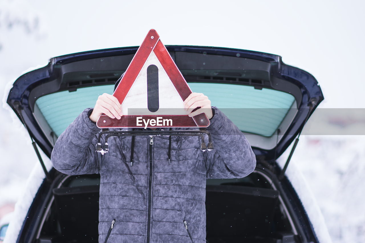 Man wearing warm clothing holding warning sign against car during winter