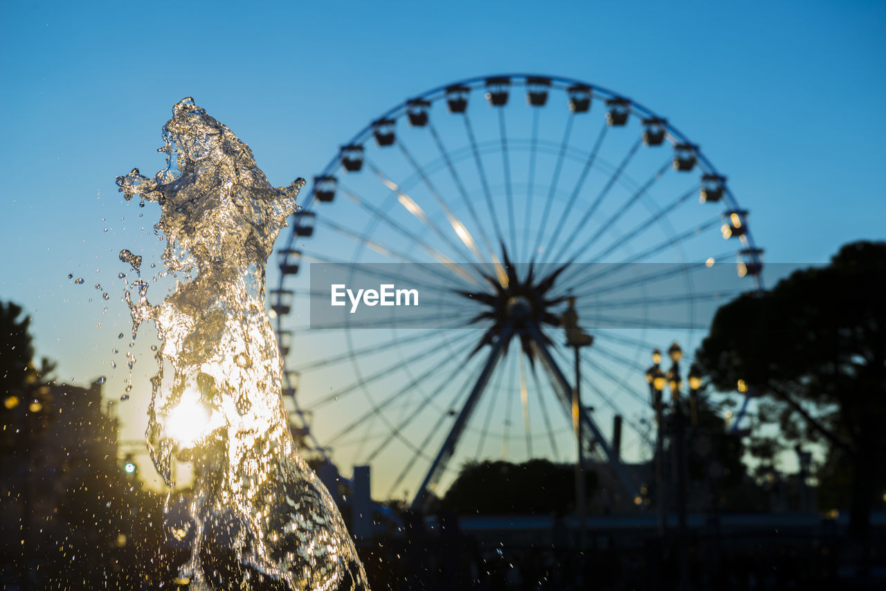 Water splashing against ferris wheel in city during sunset