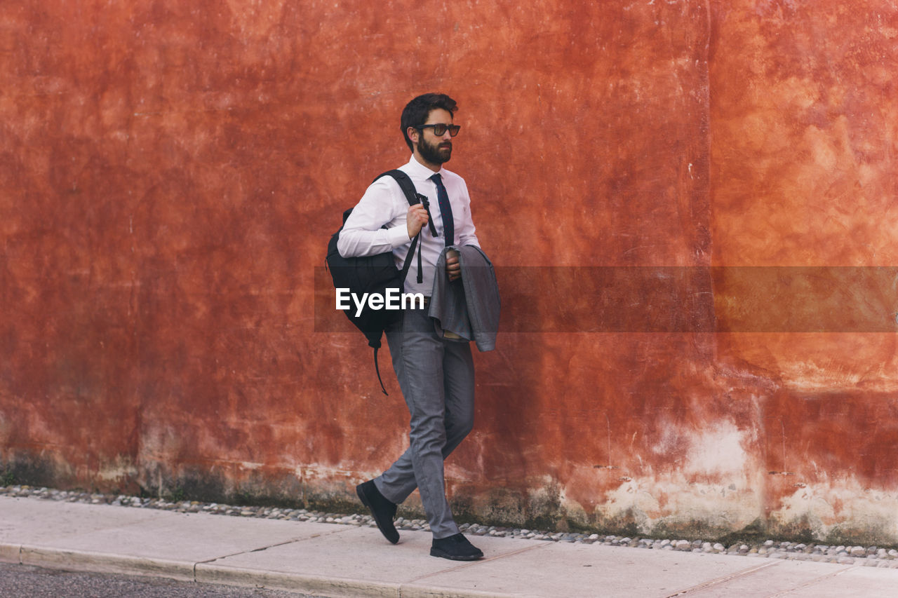 Full length of businessman with backpack walking on sidewalk against wall in city