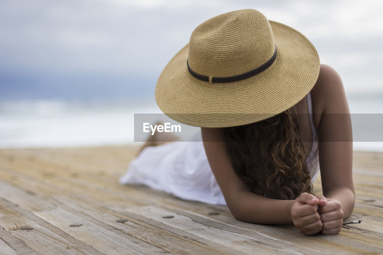 MIDSECTION OF WOMAN WEARING HAT SITTING ON BEACH