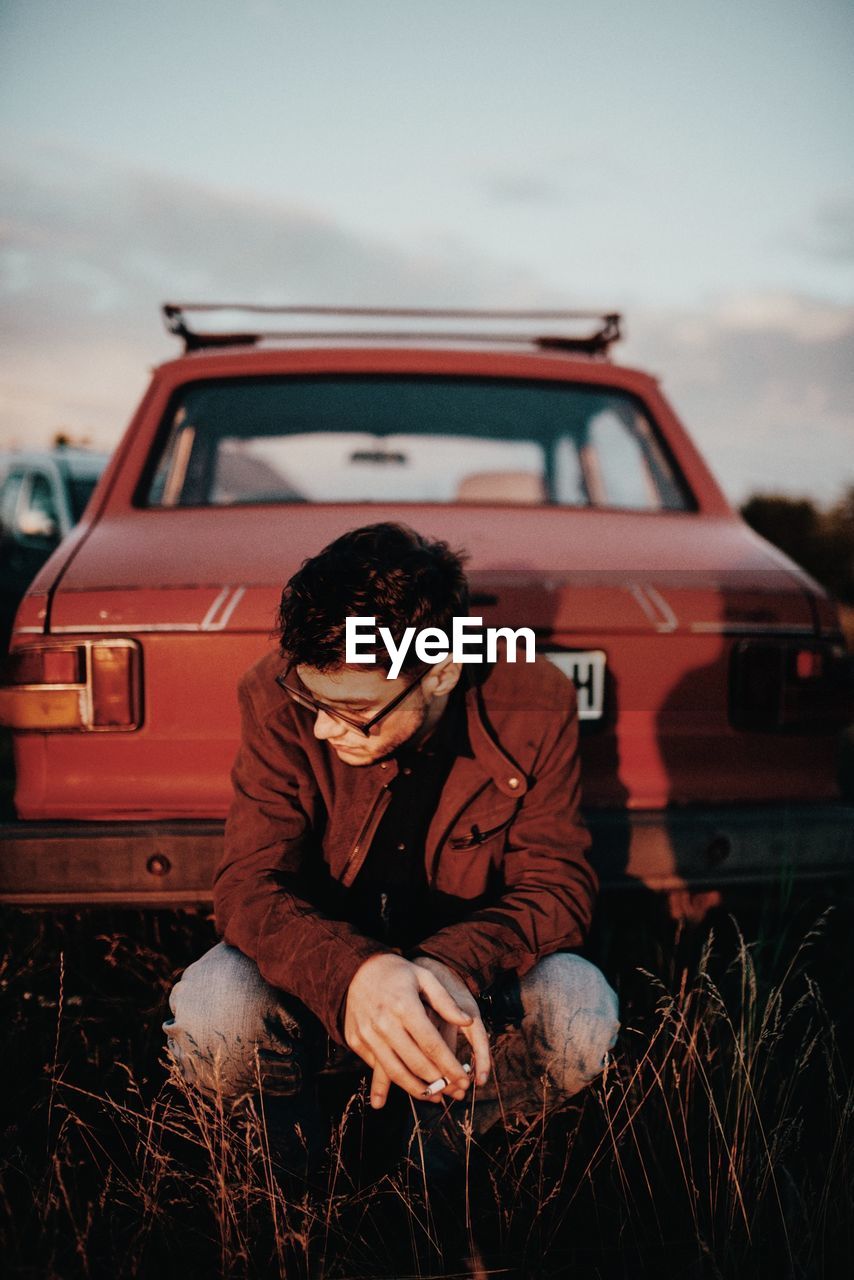 Fashionable man smoking while sitting by car on field against sky