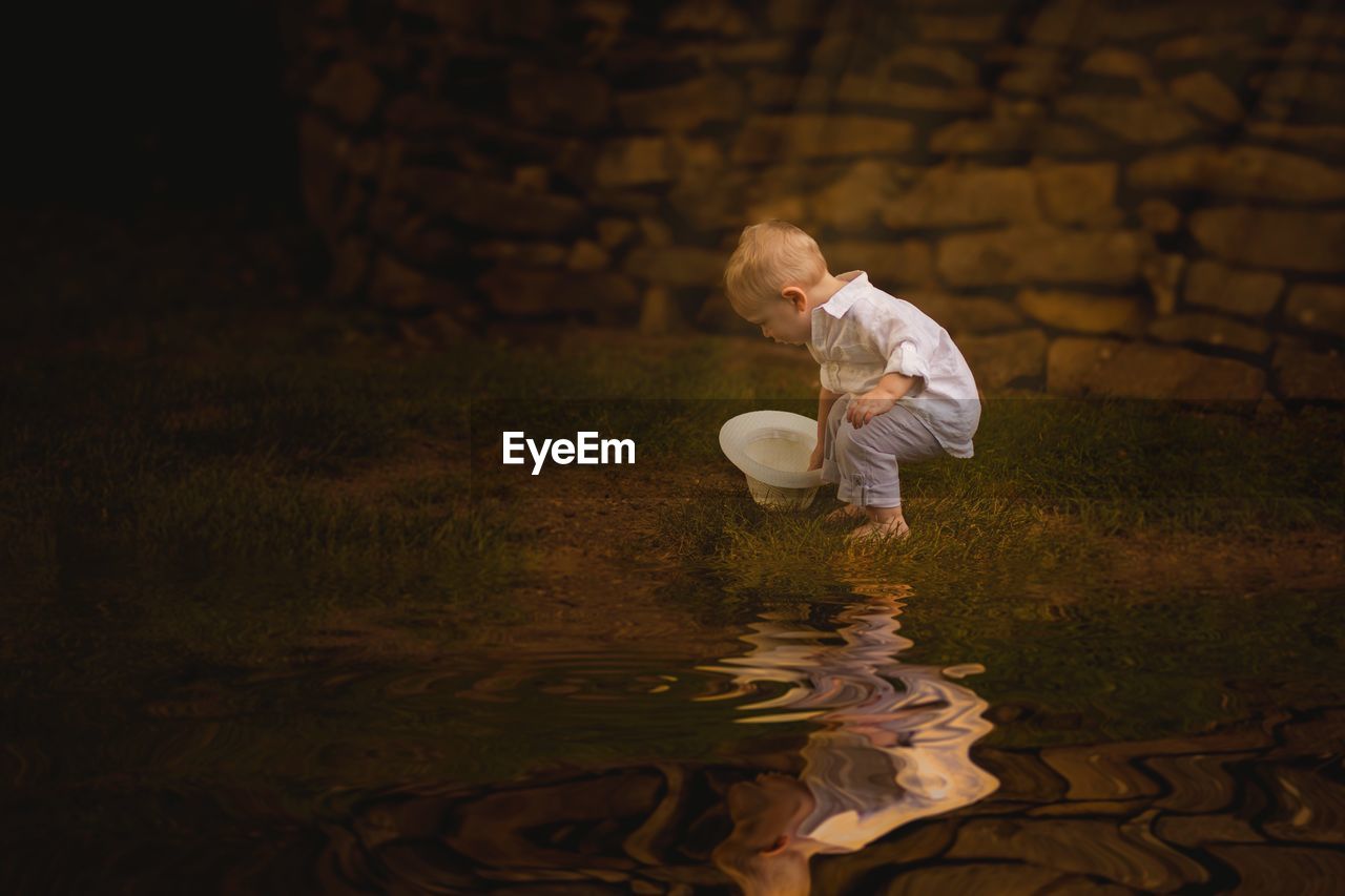 Full length side view of boy holding hat with reflection in water