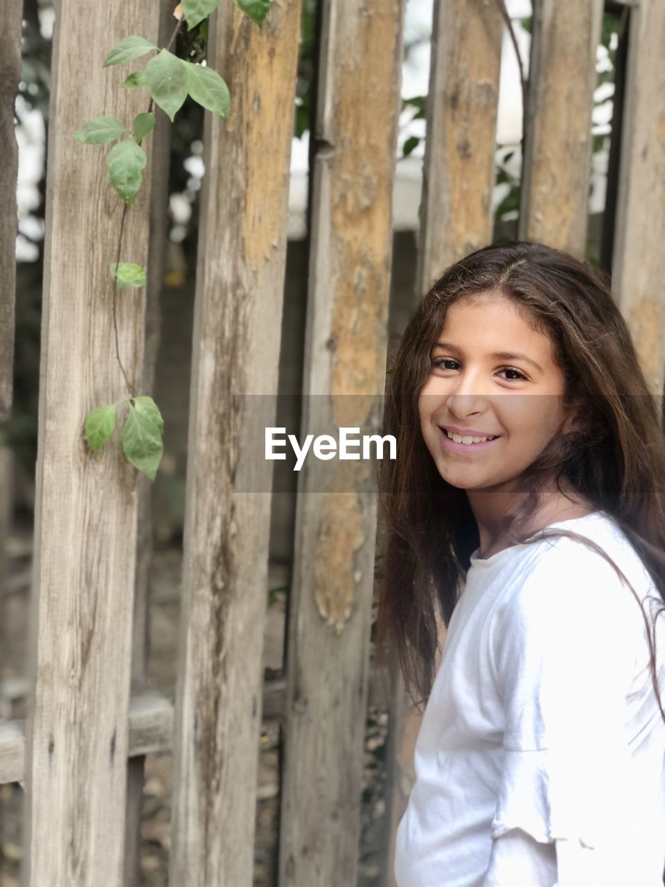 Portrait of smiling young woman standing by wooden picket fence