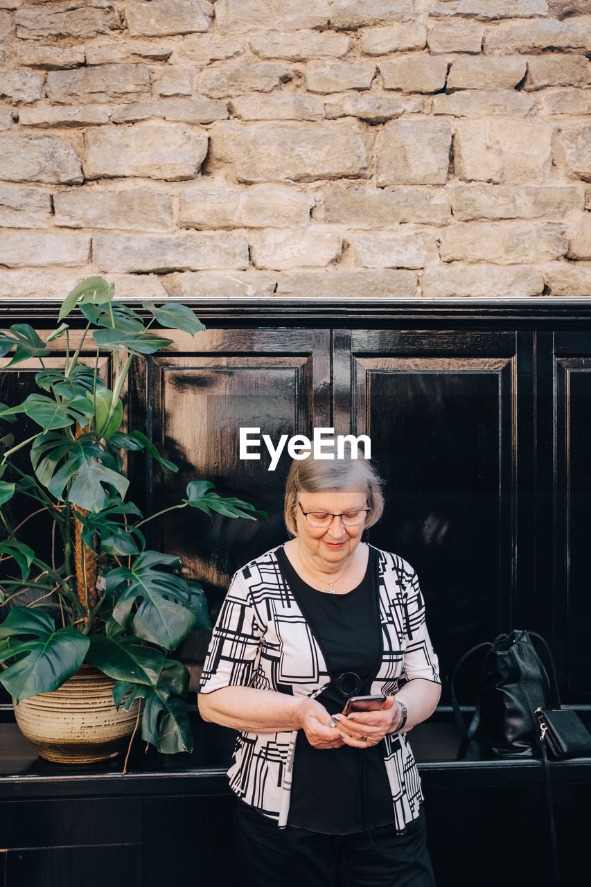 Senior female tourist using smart phone while standing against stone wall at restaurant