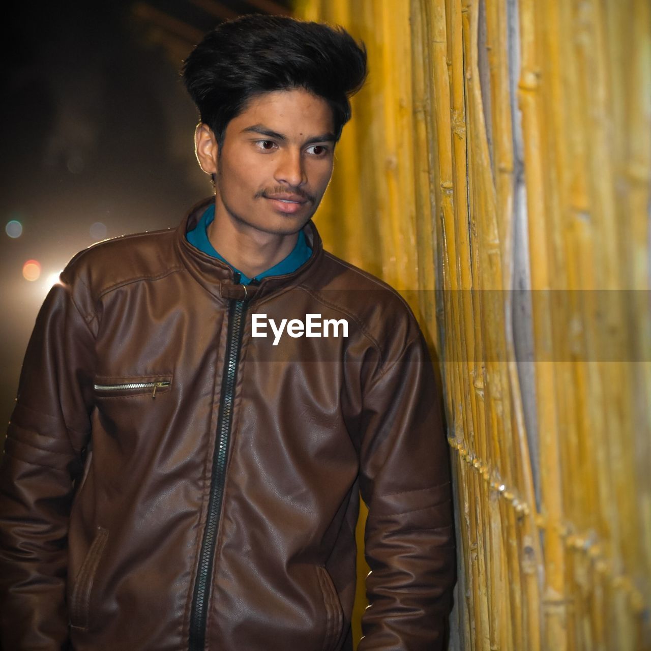 Young man looking away while standing against fence at night