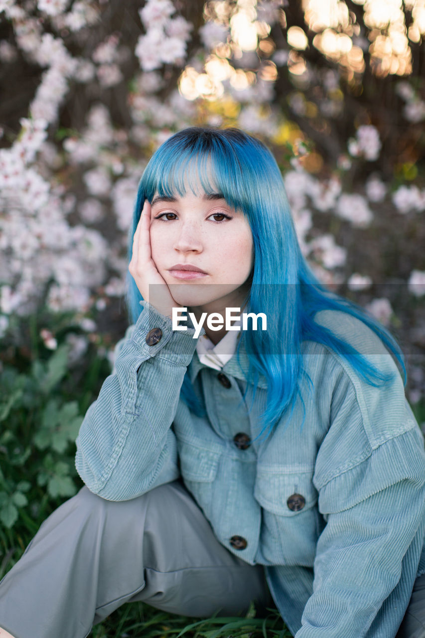 Sad millennial female model with blue hair in stylish outfit looking at camera thoughtfully while sitting on green grass near blooming tree in spring garden