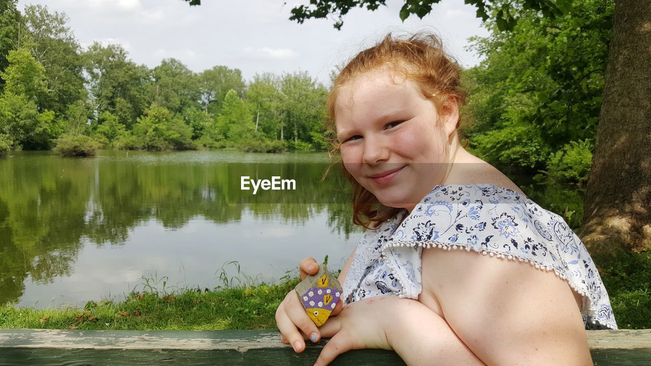 YOUNG WOMAN SITTING BY LAKE AGAINST TREES