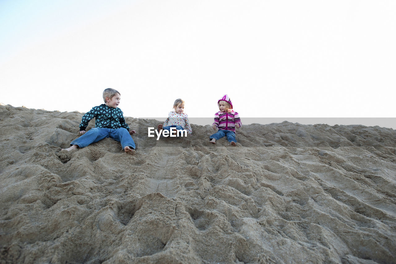 Siblings sitting on sand dune at beach against sky