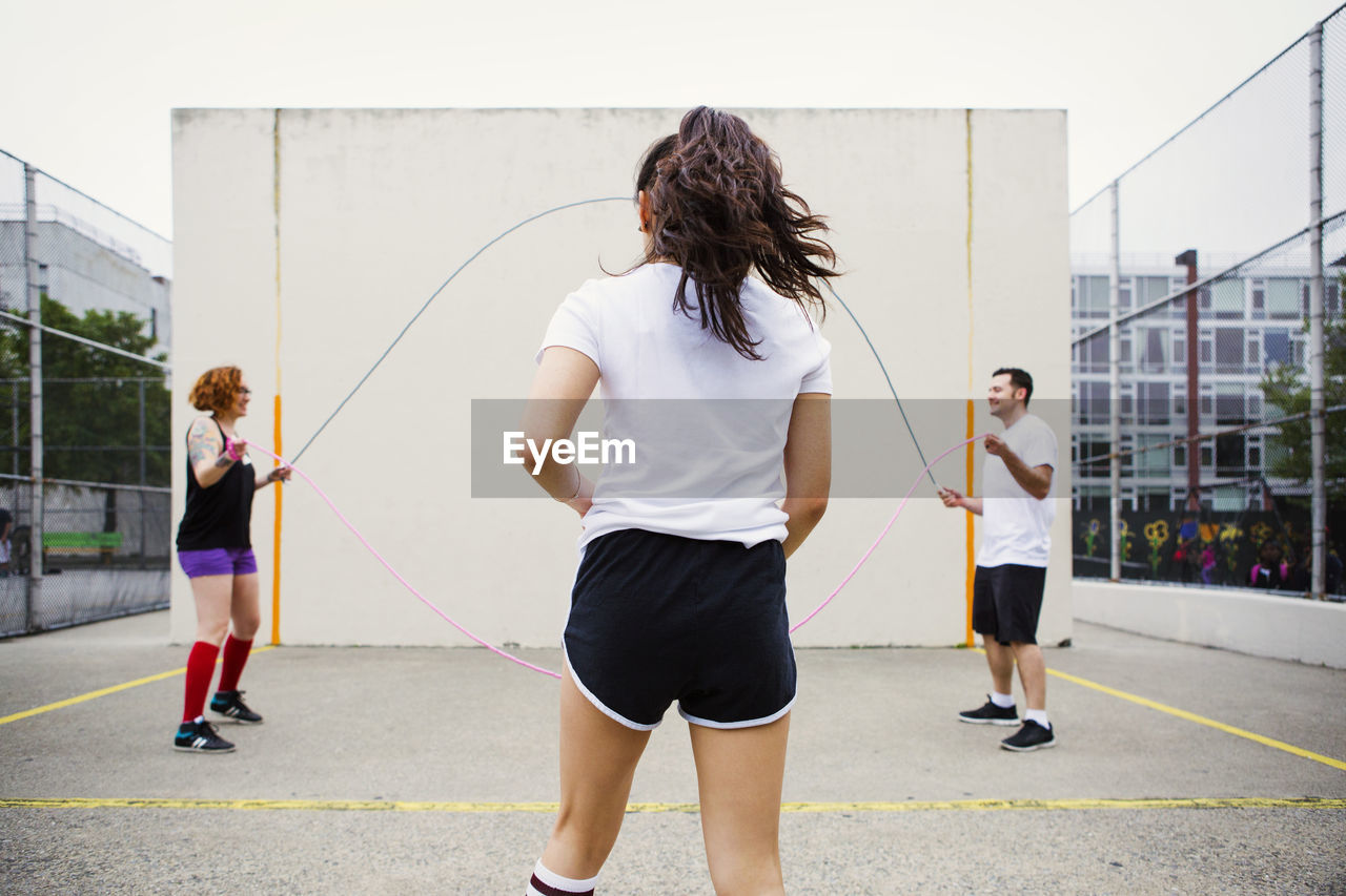 Woman walking towards friends holding jump ropes on street against wall