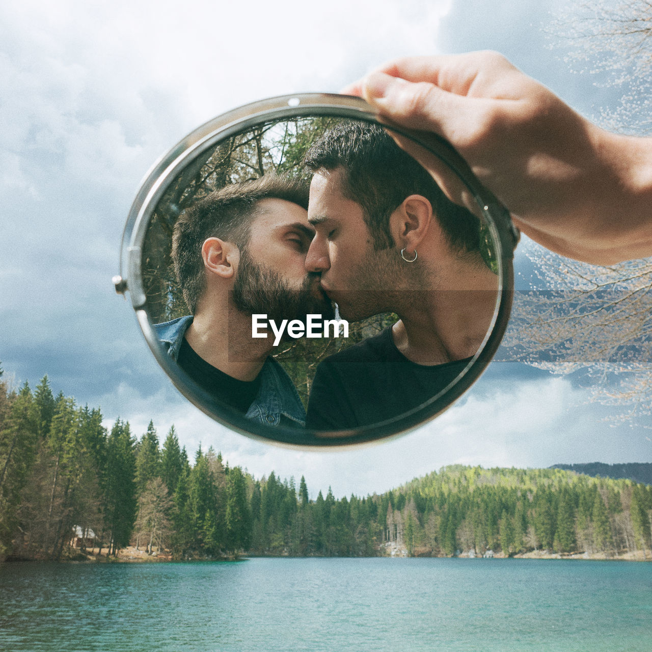 Reflection of gay couple kissing in mirror held by hand over lake against sky