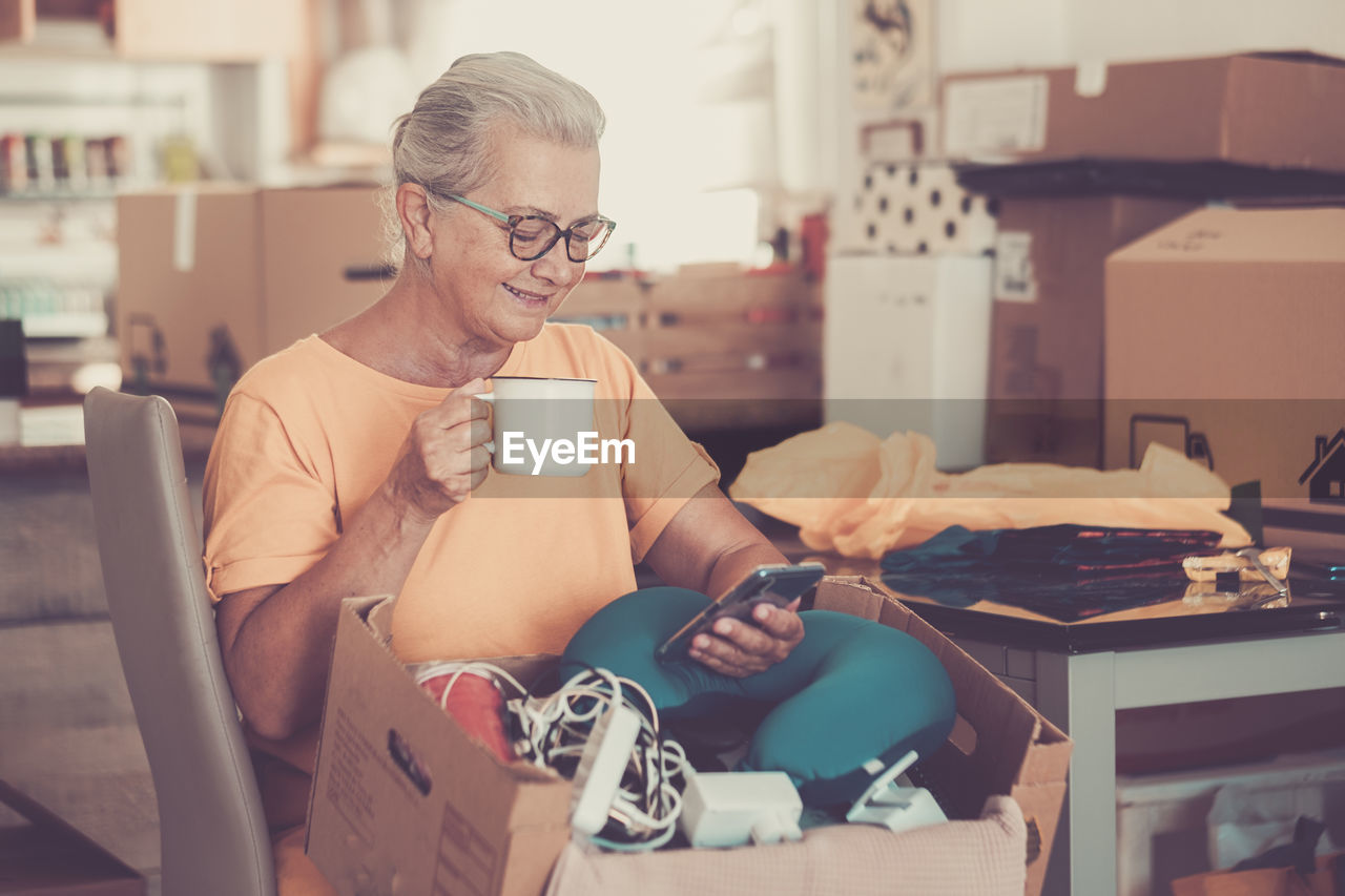 Smiling senior woman holding coffee cup using phone at home