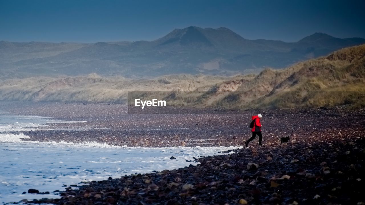 MAN STANDING ON SHORE AGAINST MOUNTAINS