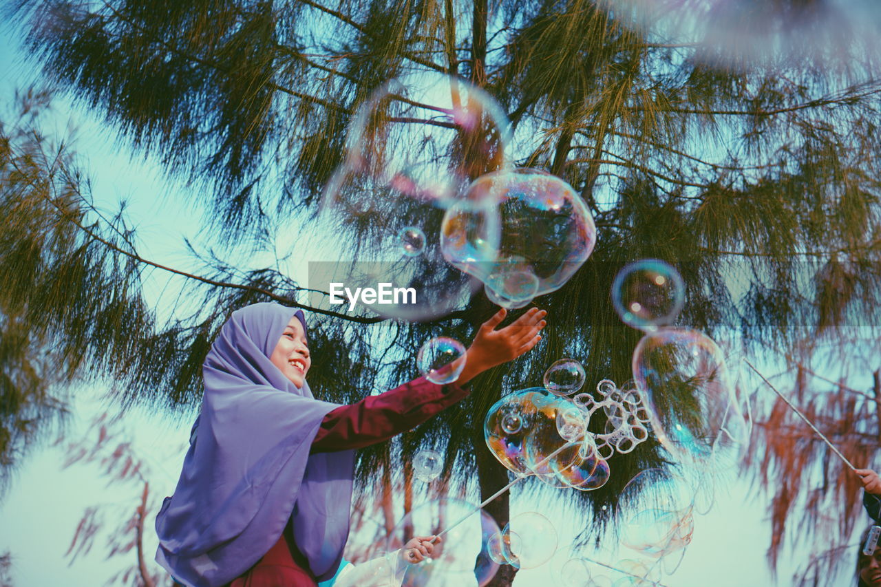 Smiling young woman in hijab playing with bubbles against tree