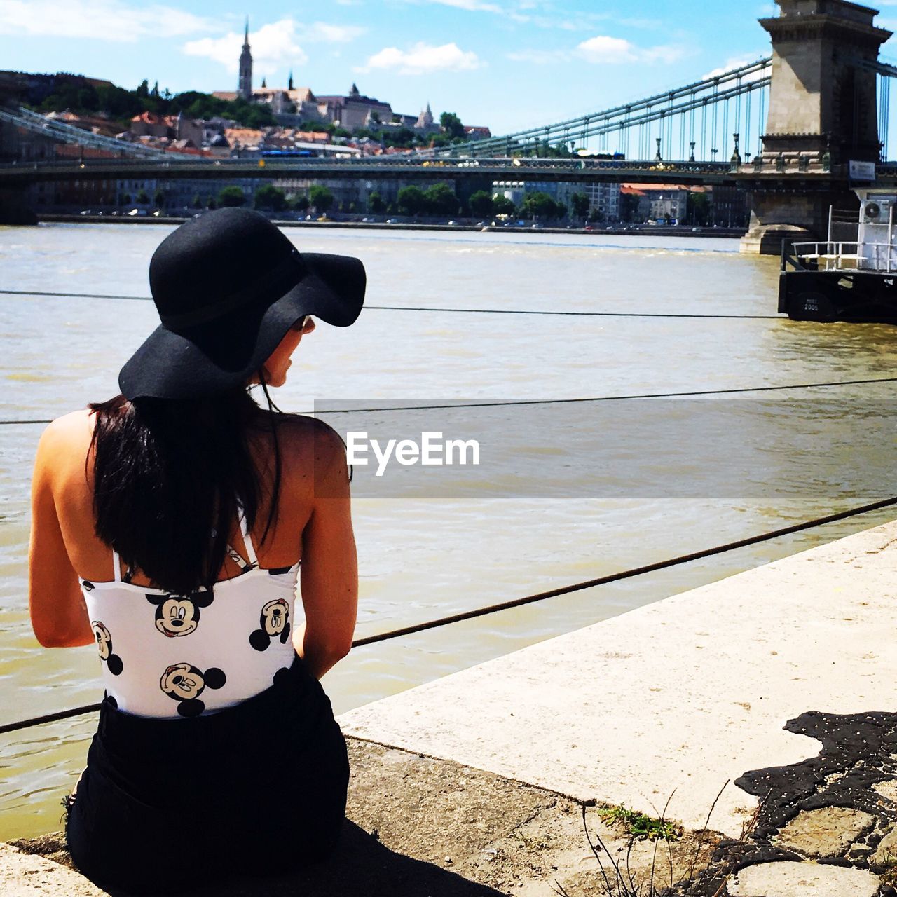 Rear view of young woman sitting on promenade against chain bridge