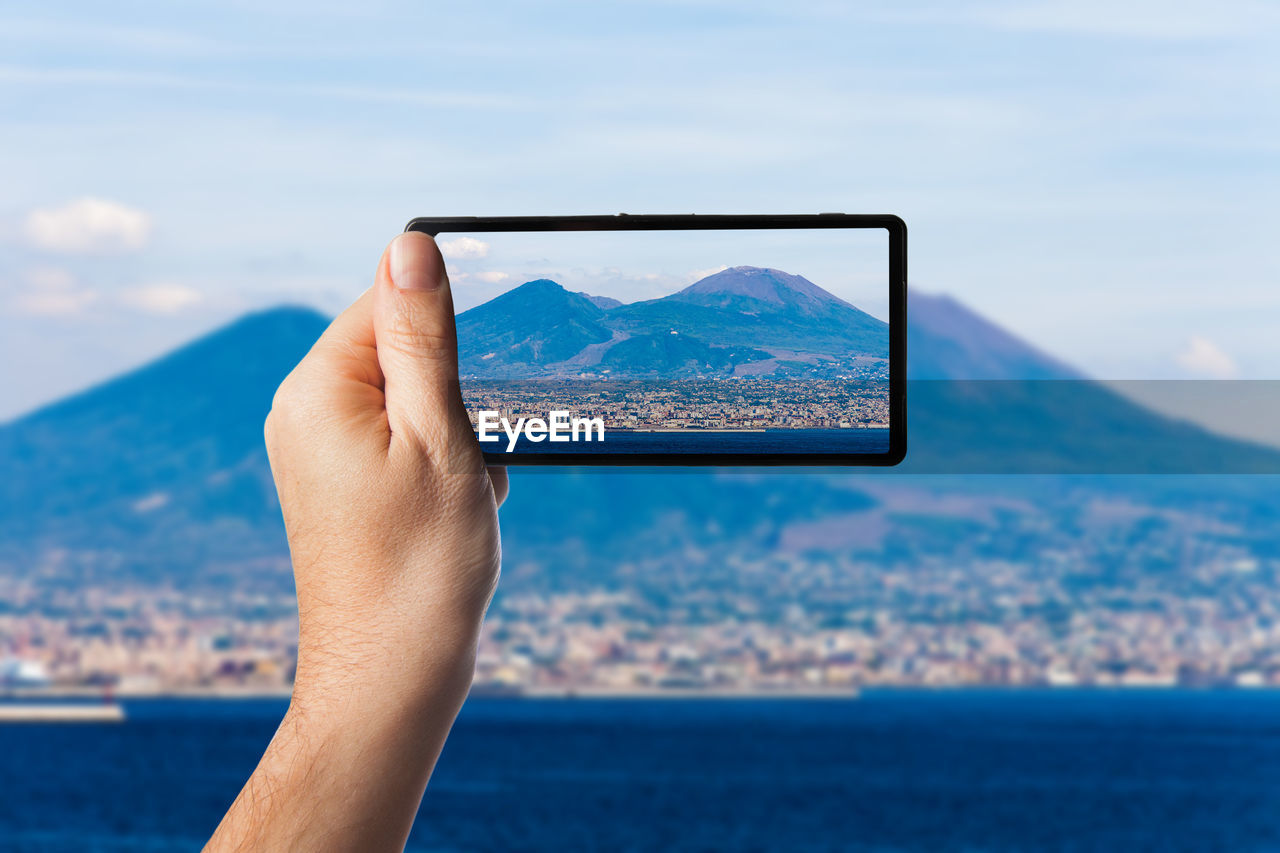 Close-up of hand photographing mountain through mobile phone against sky