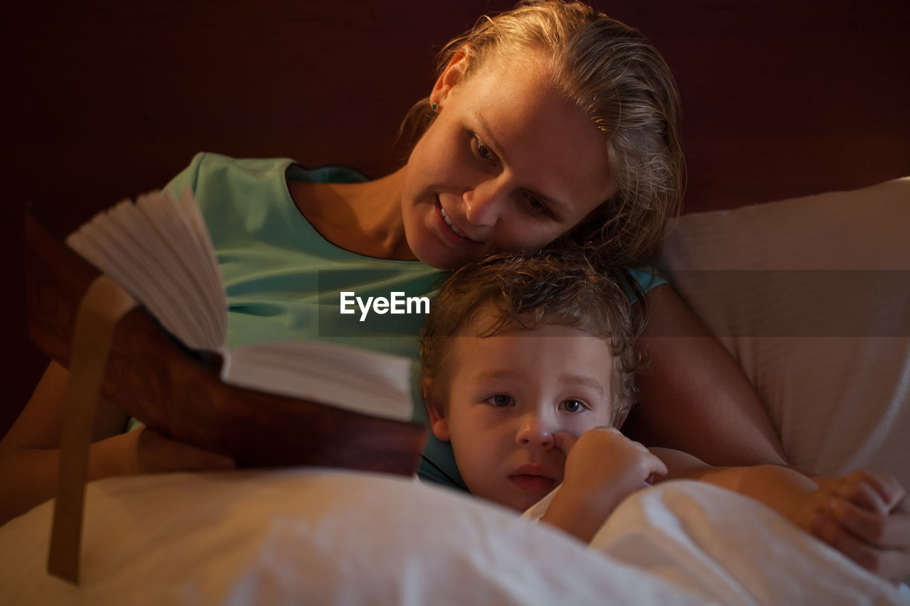 Mother holding book with son on bed in bedroom at home
