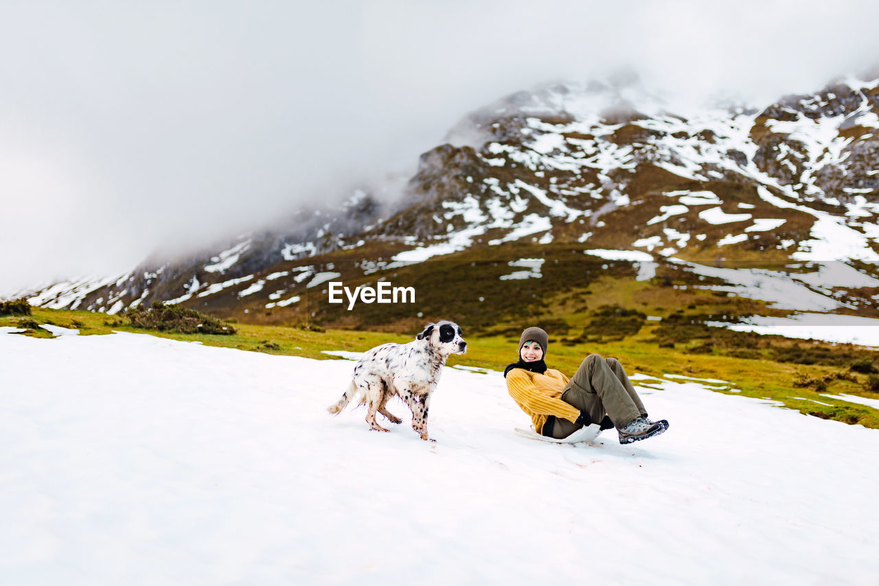 Happy woman sliding down slope covered with snow while happy english setter dog running aside in mountains