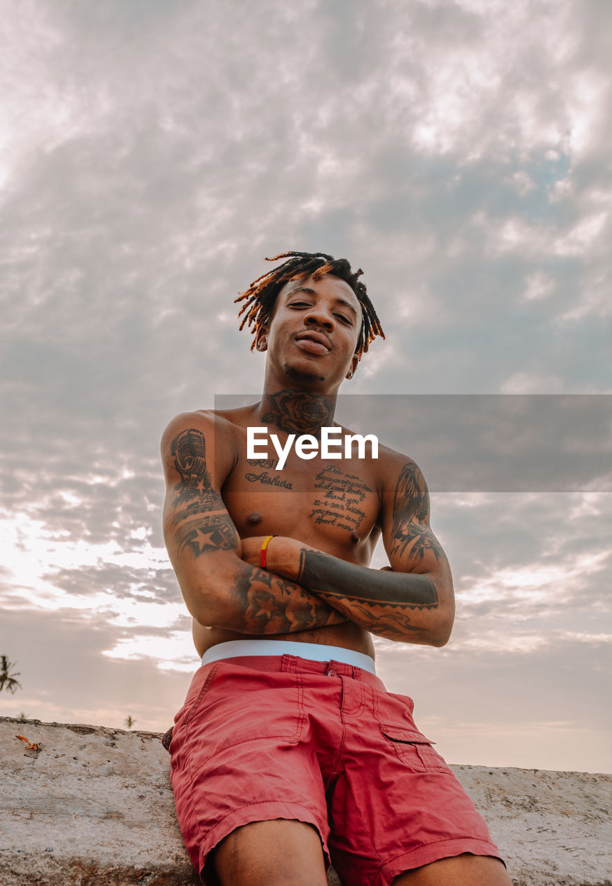 Low angle view of shirtless man with tattoos standing against sky