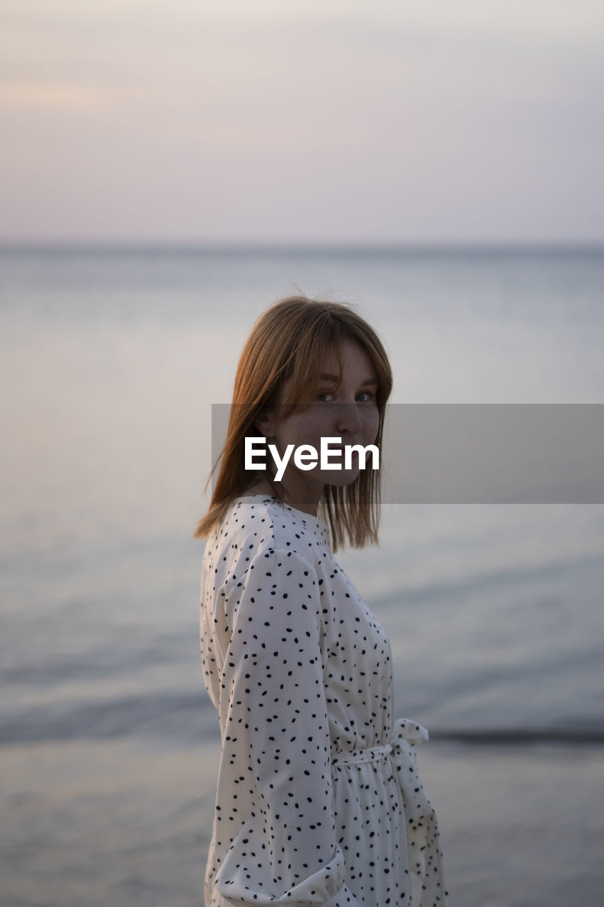 Young woman standing at beach against sky during sunset