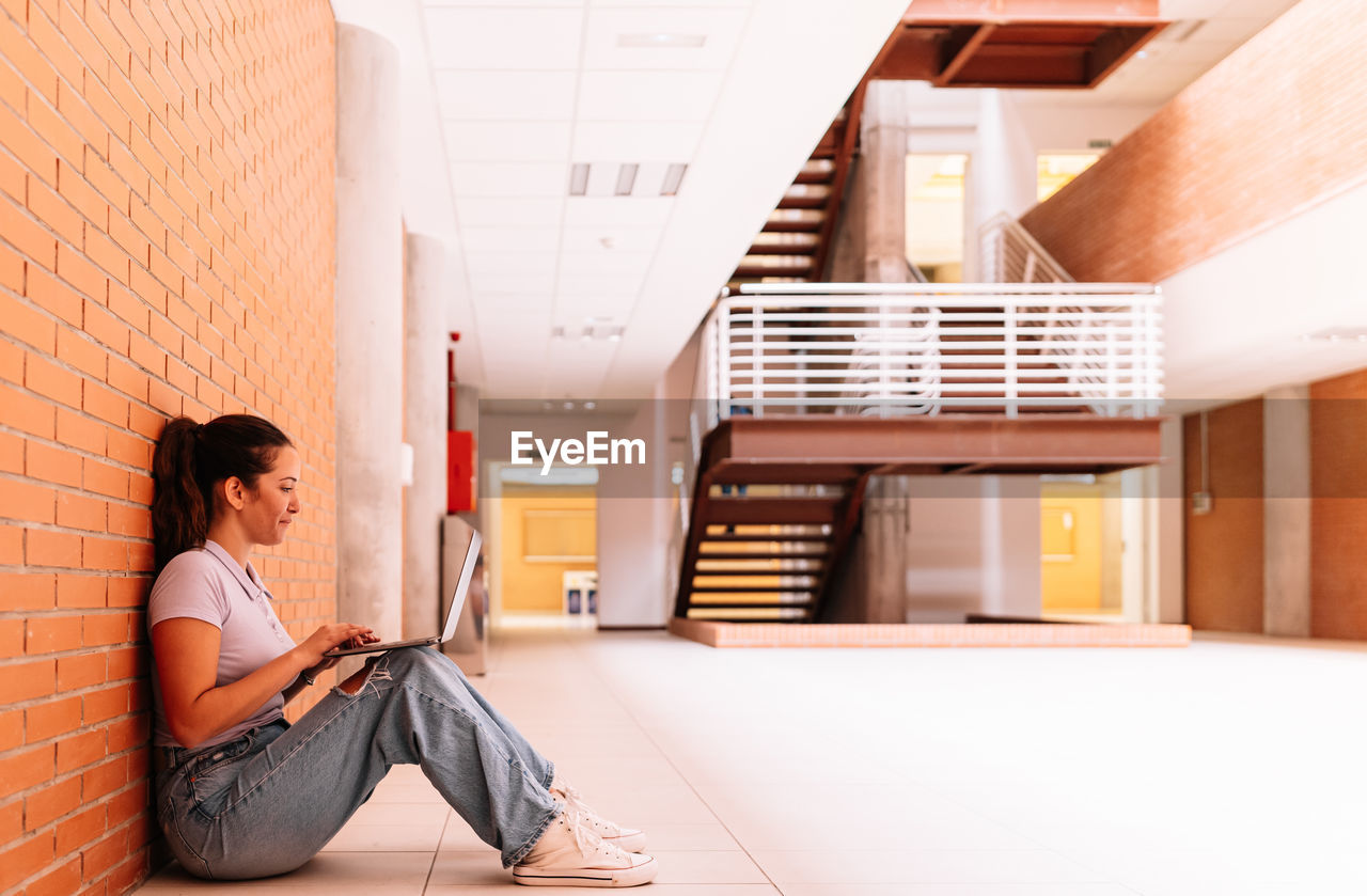 Full body side view of delighted female student in casual wear surfing internet on netbook while sitting on floor near brick wall