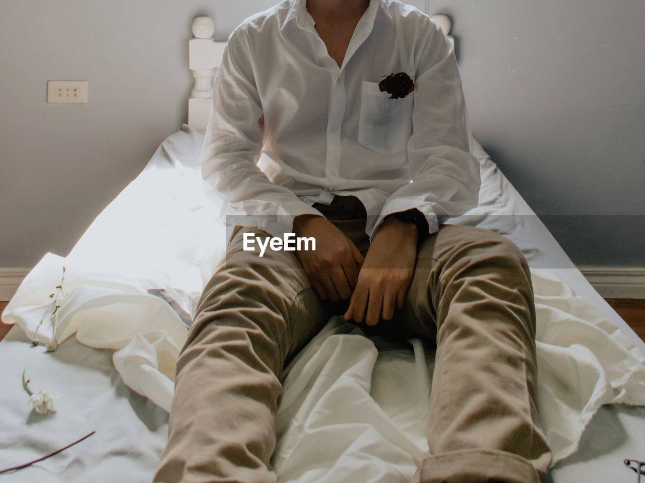 A person in white long sleeves with a rose on its pocket and khaki pants sitting in bed, alone in an 