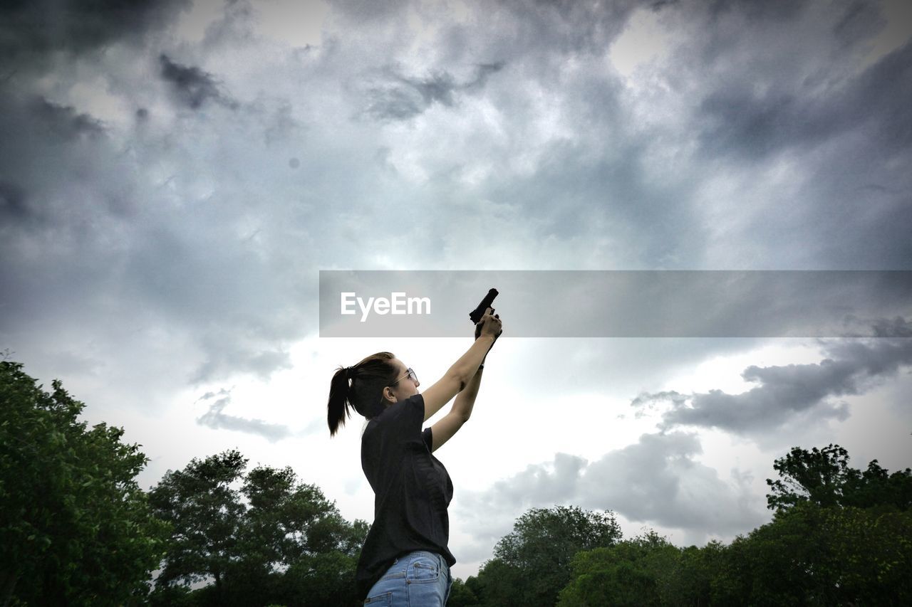 Low angle view of woman aiming with gun towards stormy clouds