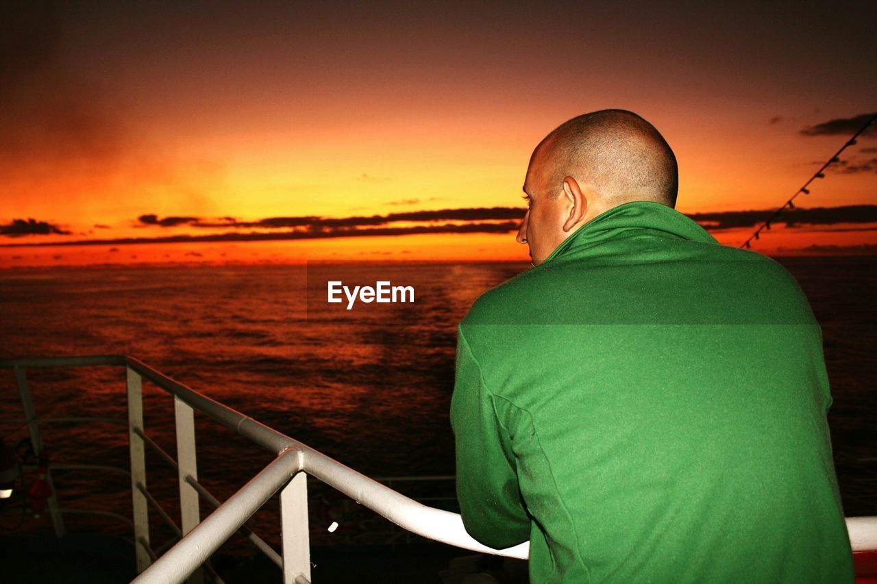 Rear view of man leaning on railing while looking at sunset