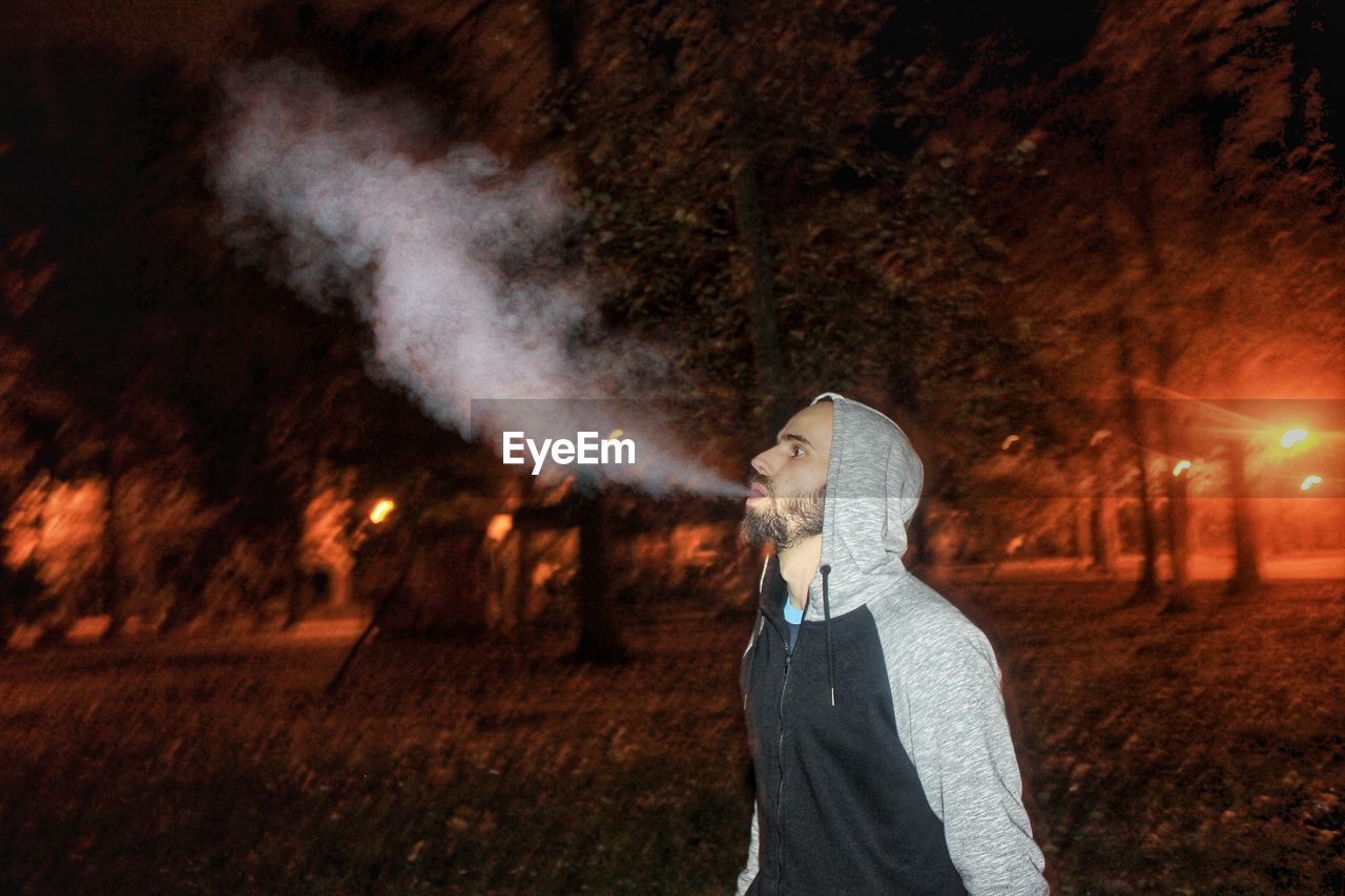 Man wearing hooded shirt while blowing smoke in park at night