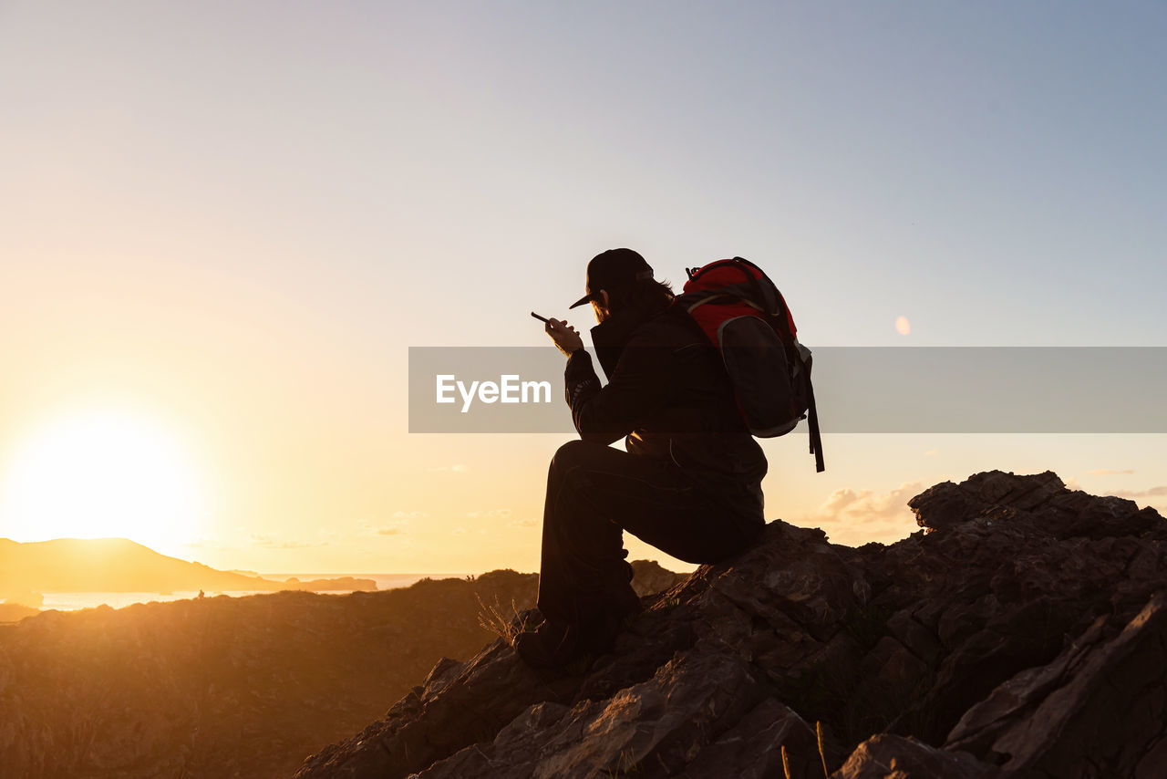 Side view of unrecognizable male hiker sitting on rock in highlands and speaking on cellphone while admiring sunset