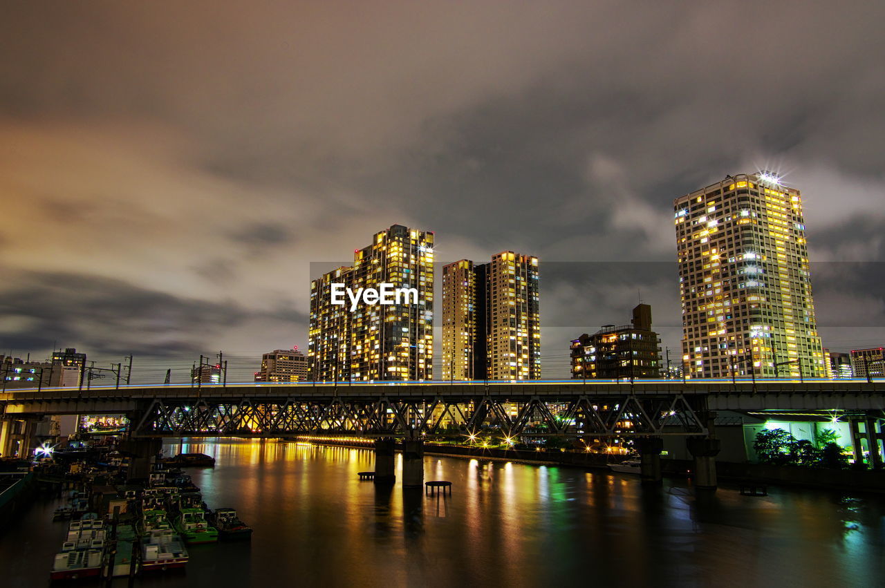 Illuminated modern buildings by river against sky at night