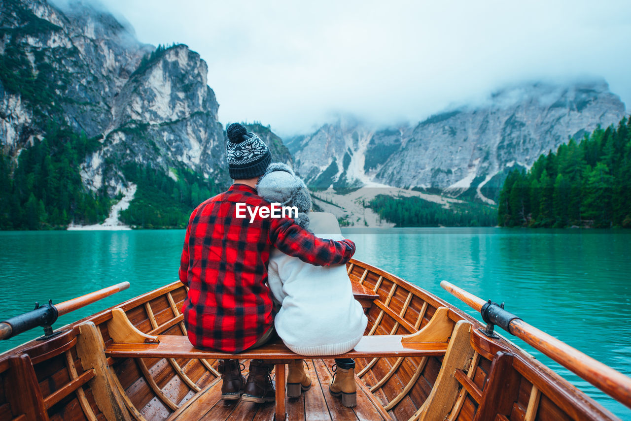 REAR VIEW OF MAN SITTING ON BOAT AT LAKE