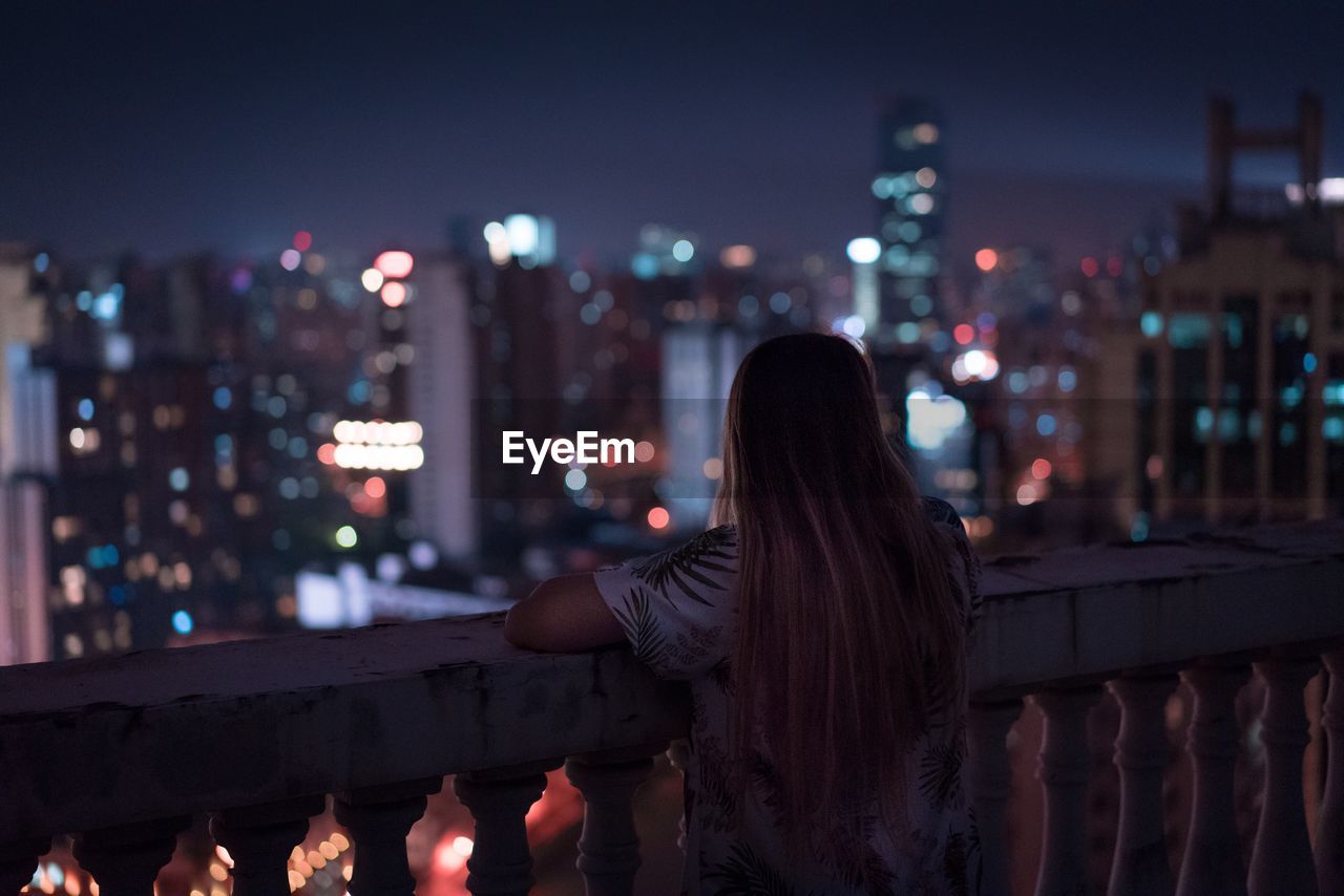 REAR VIEW OF WOMAN WITH ILLUMINATED CITYSCAPE AGAINST SKY