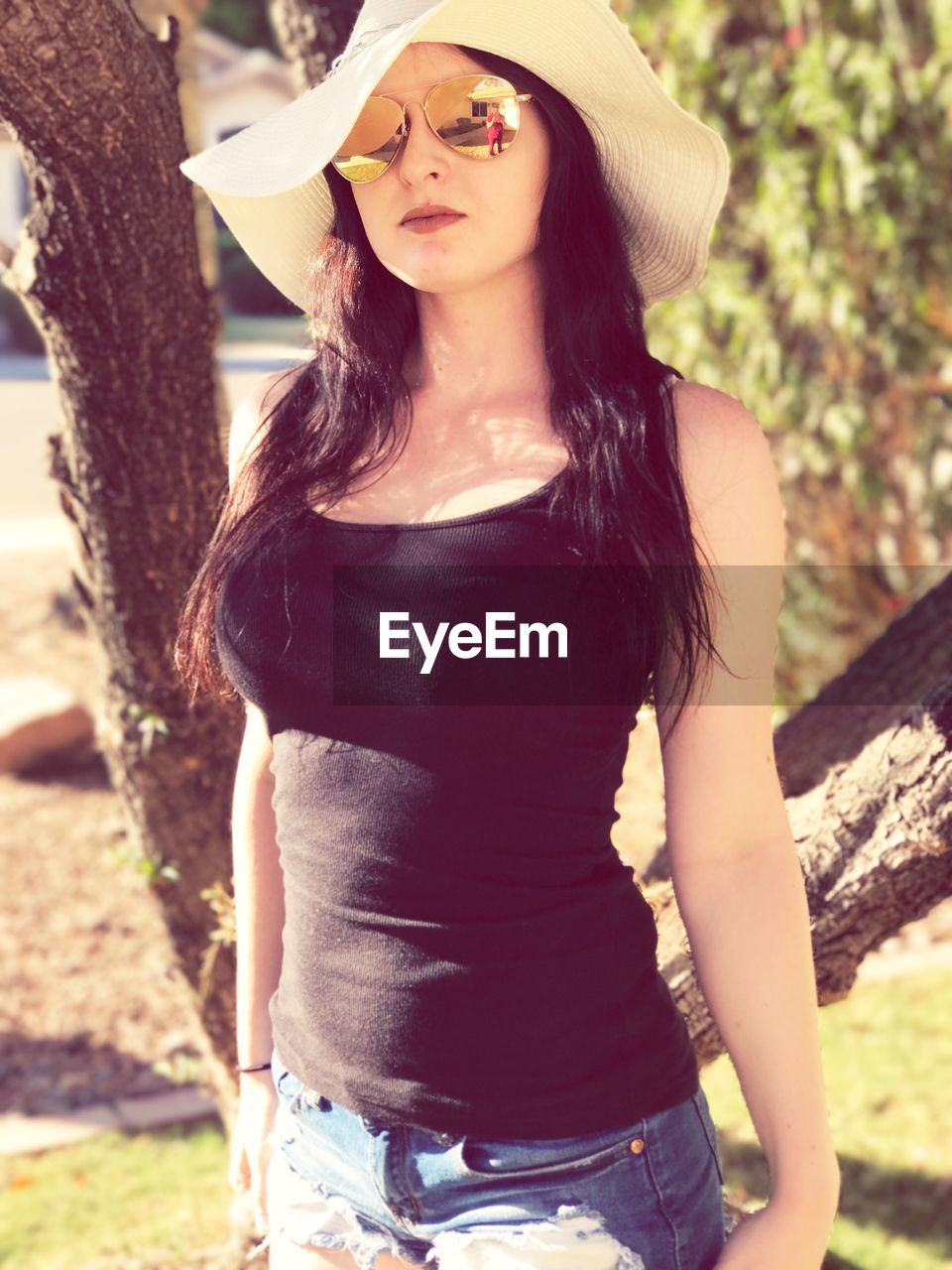 Portrait of young woman wearing sun hat and sunglasses while standing against tree at park