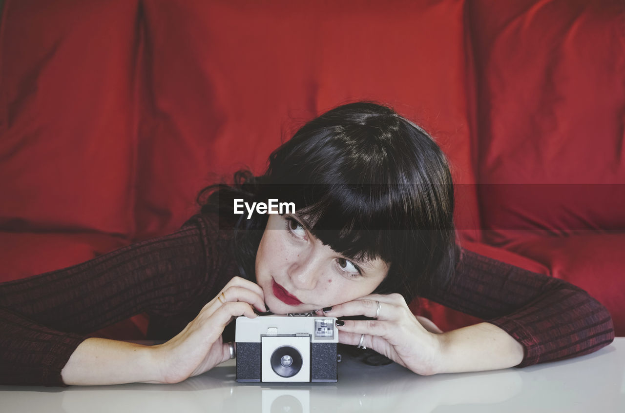 Thoughtful woman leaning on analog camera in front of red couch