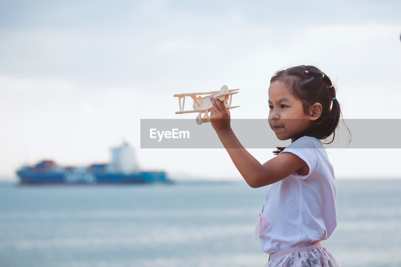 Side view of girl holding toy airplane at beach against sky