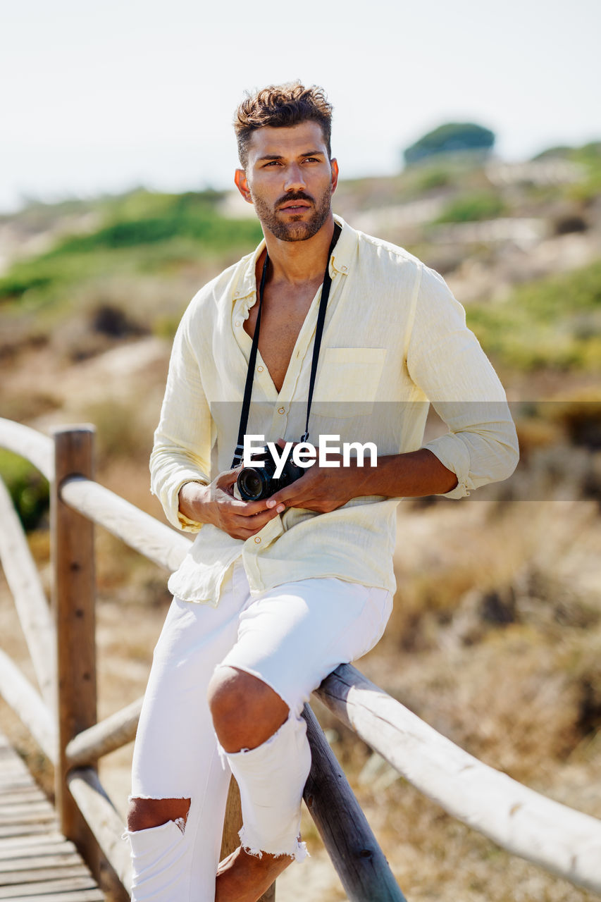 Young man holding camera sitting on railing against sky