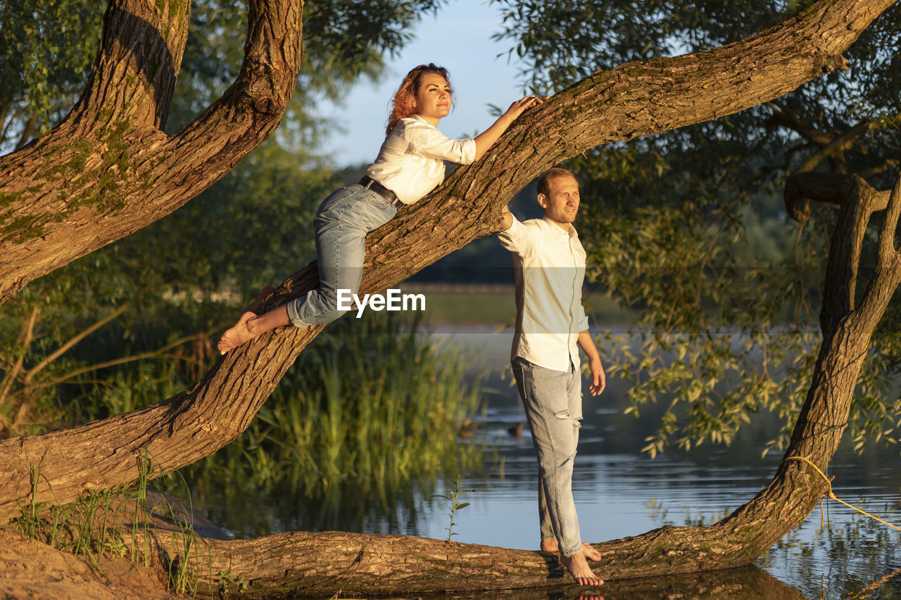 Latin woman lying on green tree branch, caucasian man stands nearby and look into distance, happy