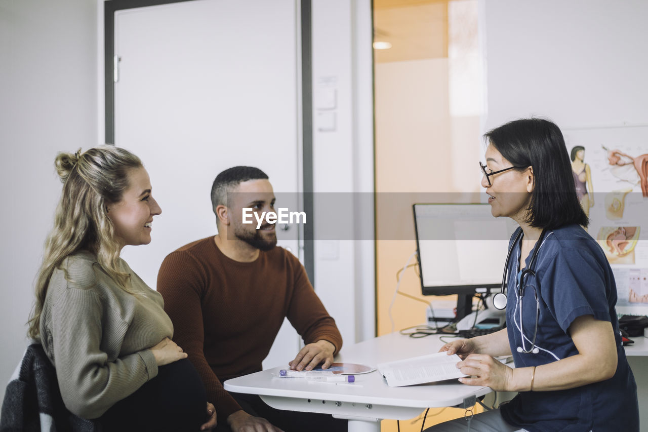 Smiling female doctor discussing with pregnant woman sitting by man at desk in clinic