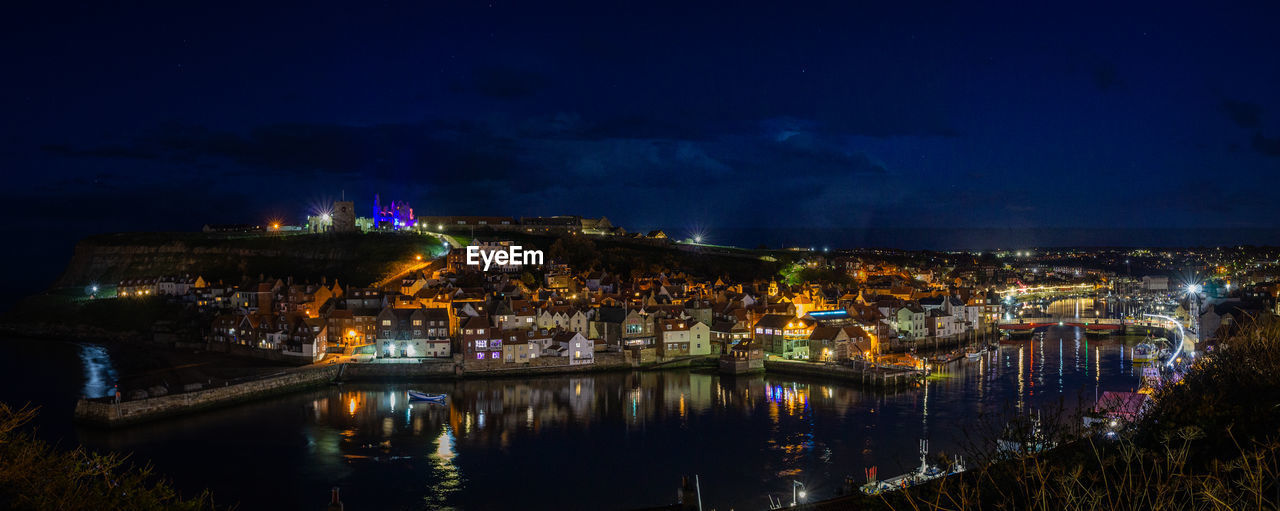 Illuminated buildings by river against sky at night