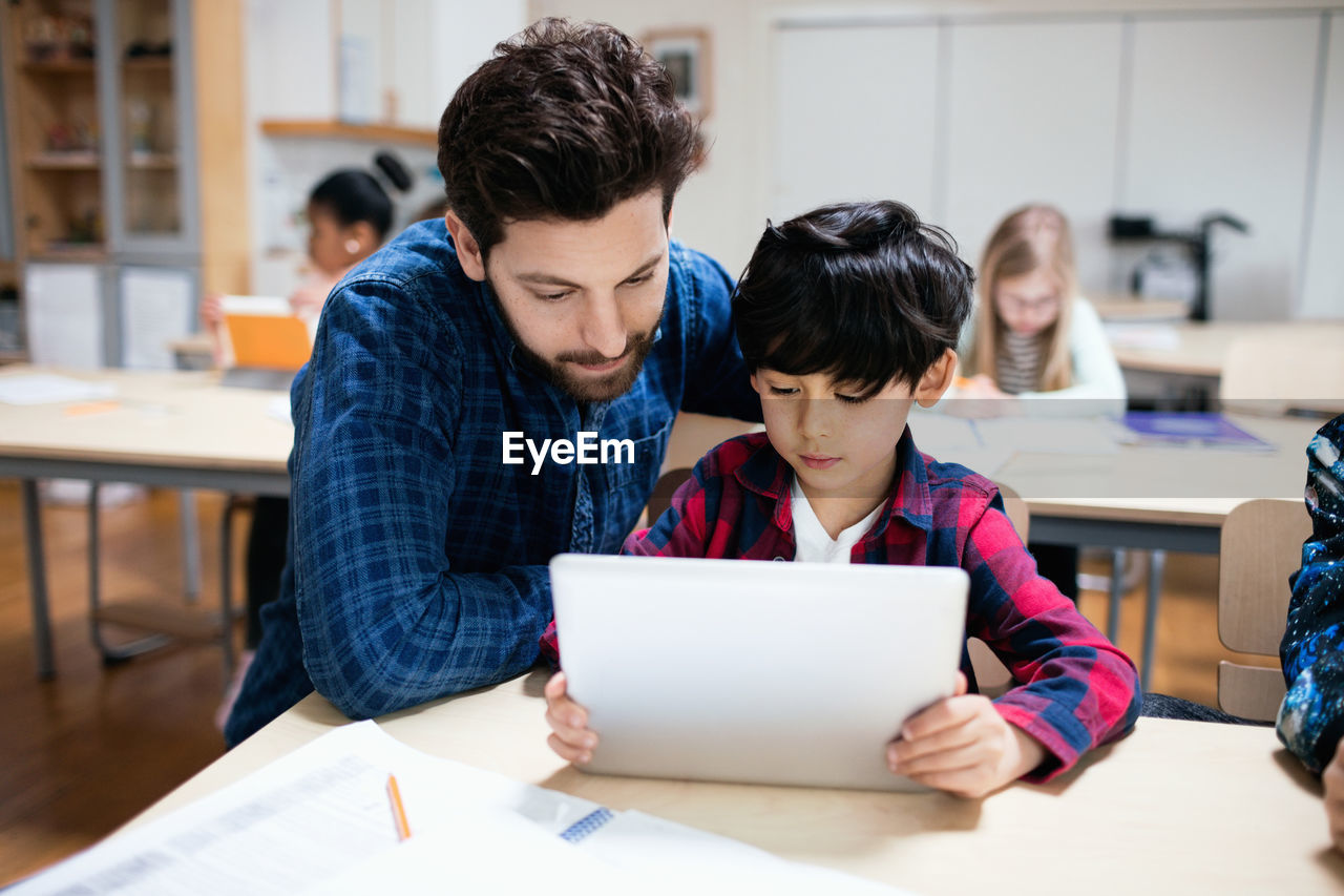 Teacher assisting male student in using digital tablet while sitting in classroom