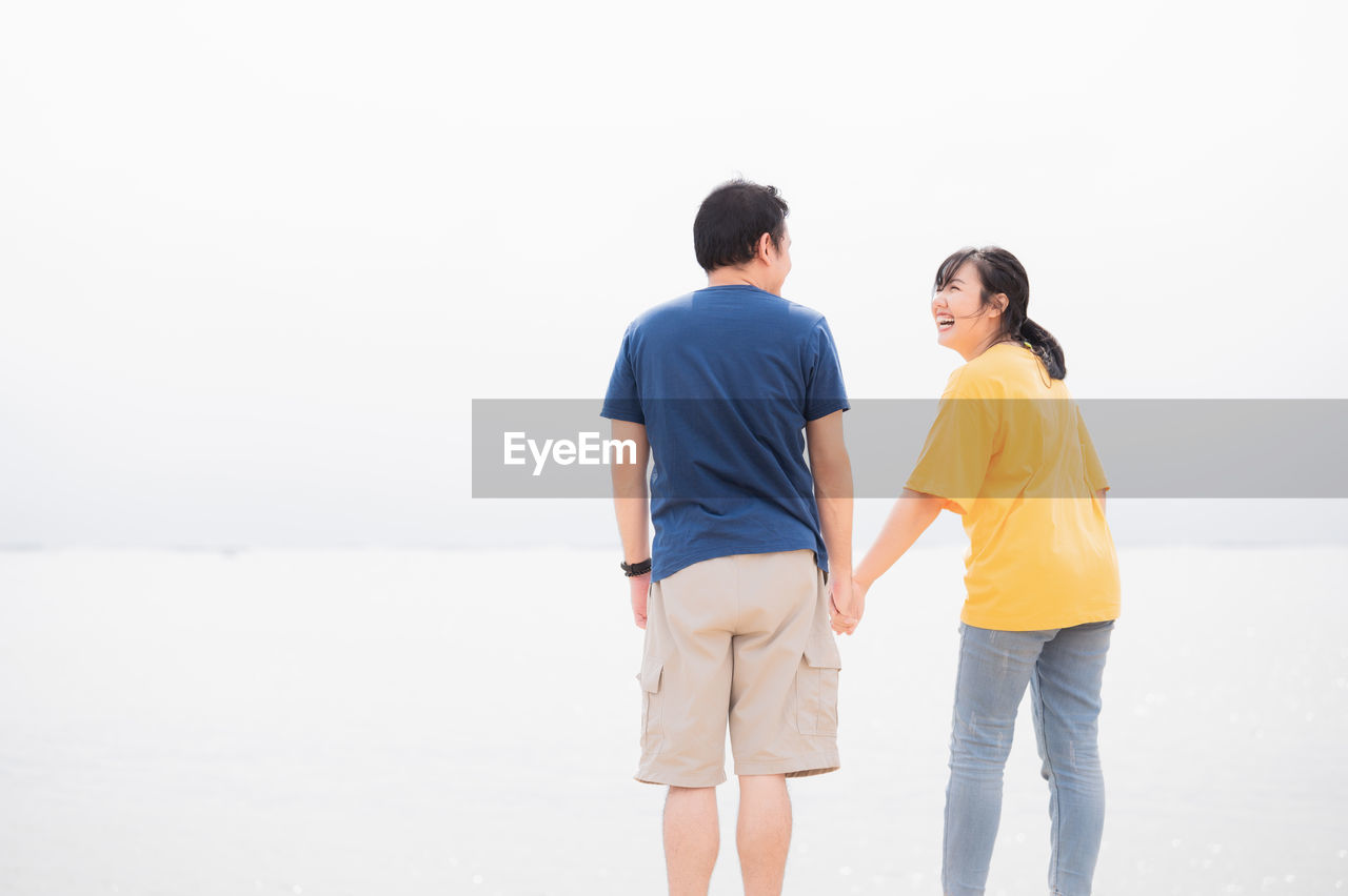 REAR VIEW OF COUPLE STANDING AGAINST WATER AT SHORE