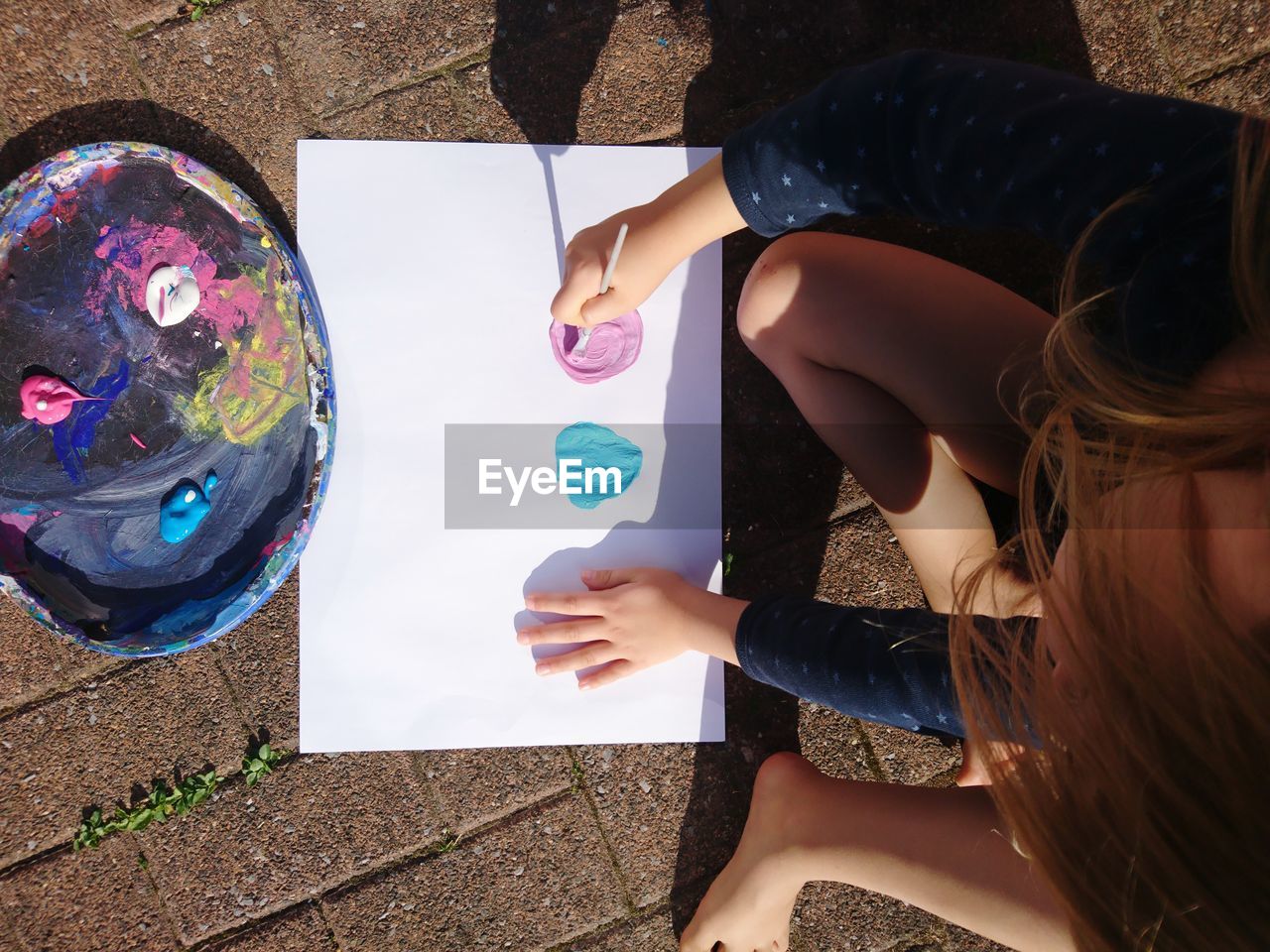 High angle view of girl painting on paper while sitting at footpath